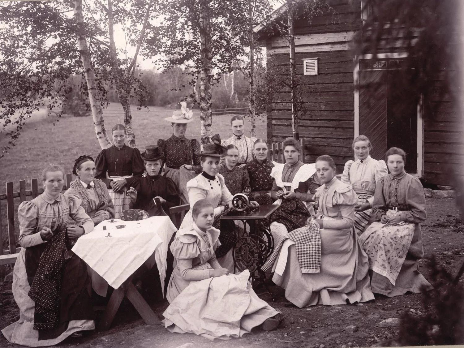 Svartvitt foto - en grupp människor sittande utomhus, med trädgård och träd i bakgrunden.