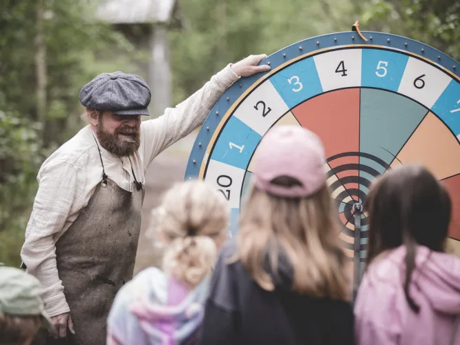 Färgbild - en grupp barn stående framför ett tombolahjul i Trolska Skogen. En sagokaraktär snurrar hjulet.
