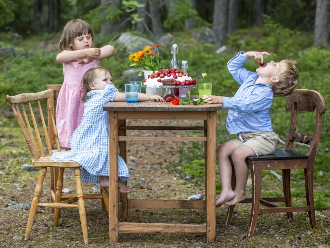 Färgbild - tre barn vid ett dukt sommarbord utomhus. Länkad bild.