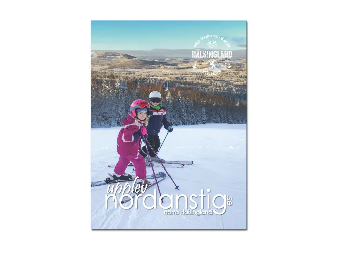 Färgbild - framsida på turistmagasin Nordanstig 2026. Bild Två barn stående i skidbacke med vinterlandskapet i bakgrunden.