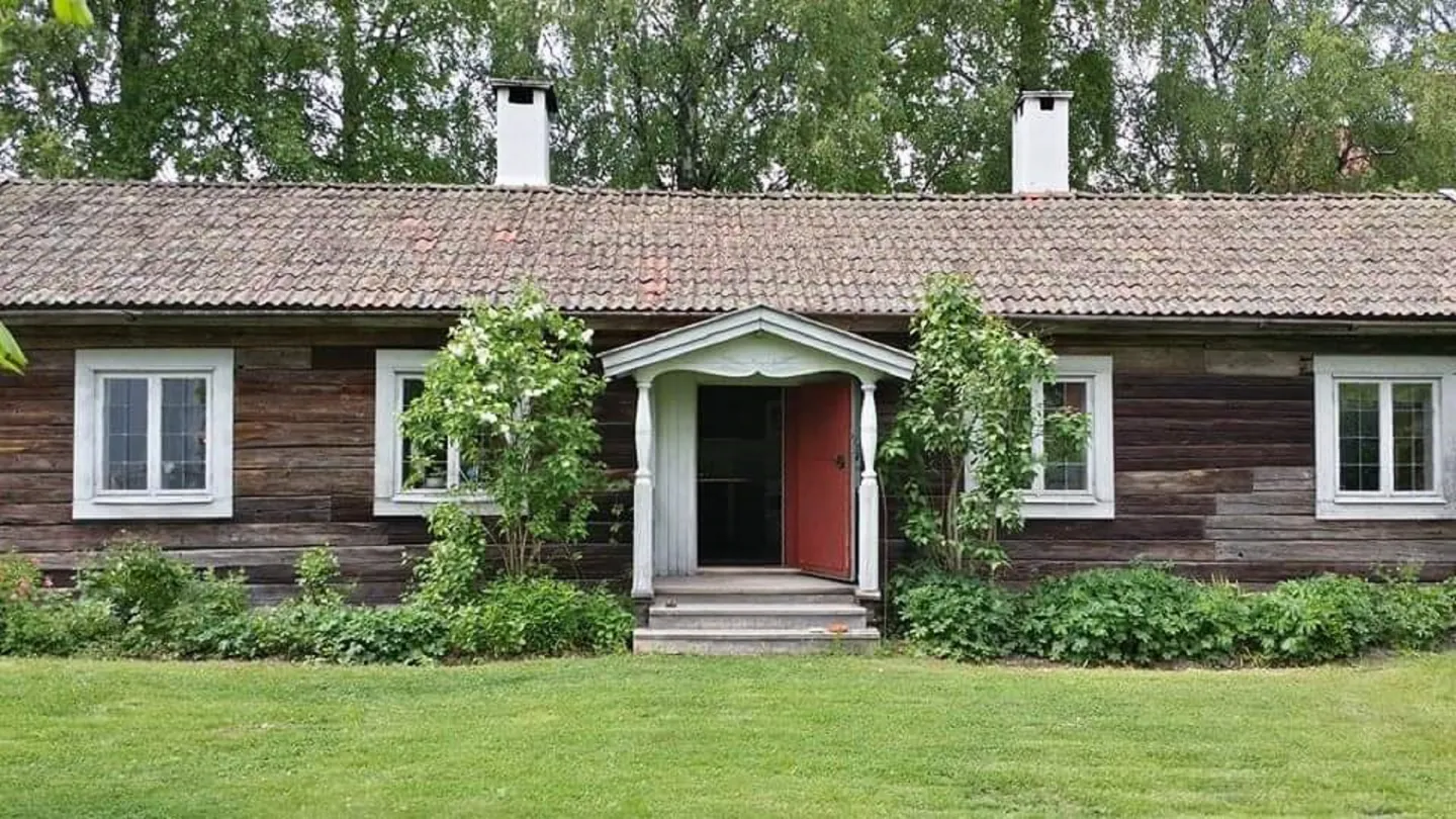 Grå timmerstuga med vita fönsterkarmar och brokvist i hälsingestil.
