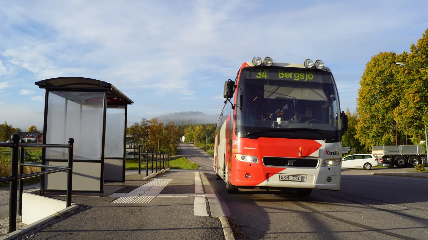 Färgbild - en X-trafikbuss vid hållplats. Omgivande sommargrönska och blå himmel.