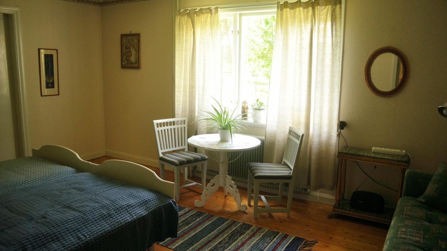 Rum med säng, bord och stolar och ett fönster med ljusa gardiner.