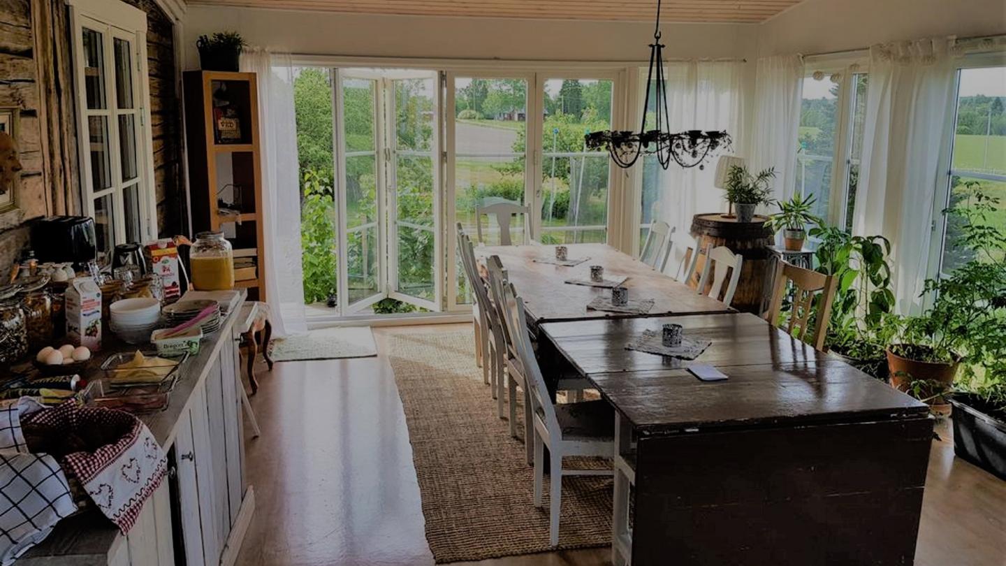 Matsalsrum med bord och stolar. Frukostbuffe uppdukad på bänk till vänster. Trädgårdsutsikt genom fönster.